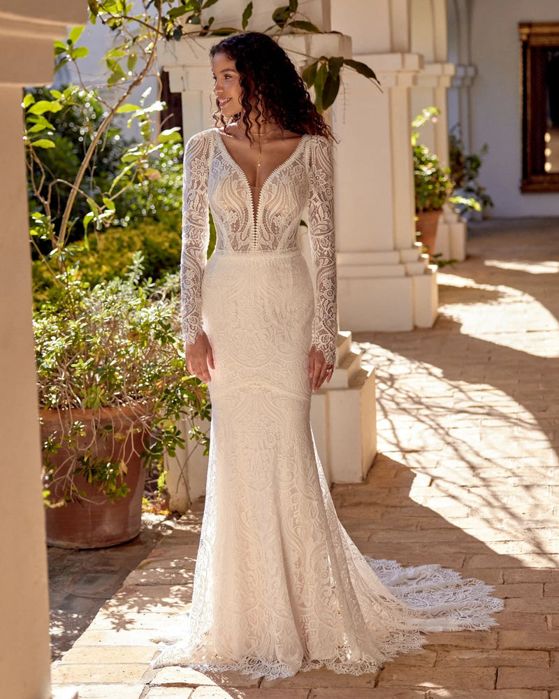 Woman in a white lace wedding dress standing on a sunlit patio with plants and arches.