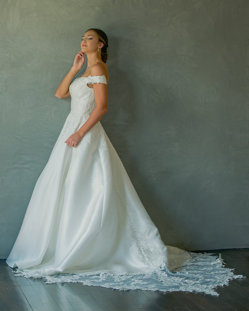 Woman in a white wedding dress standing against a gray wall with a plant on the left.