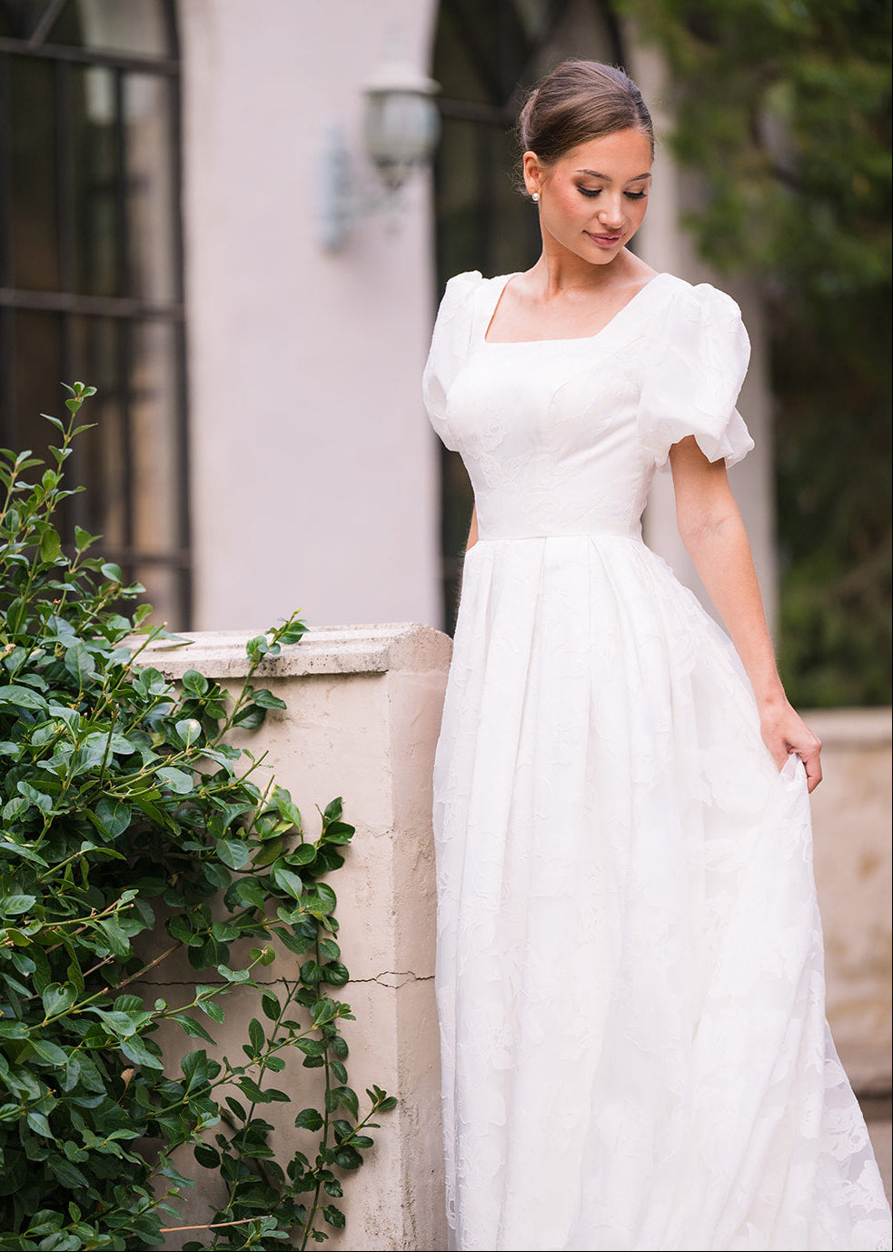Woman in a white wedding dress standing outdoors near greenery and a building.