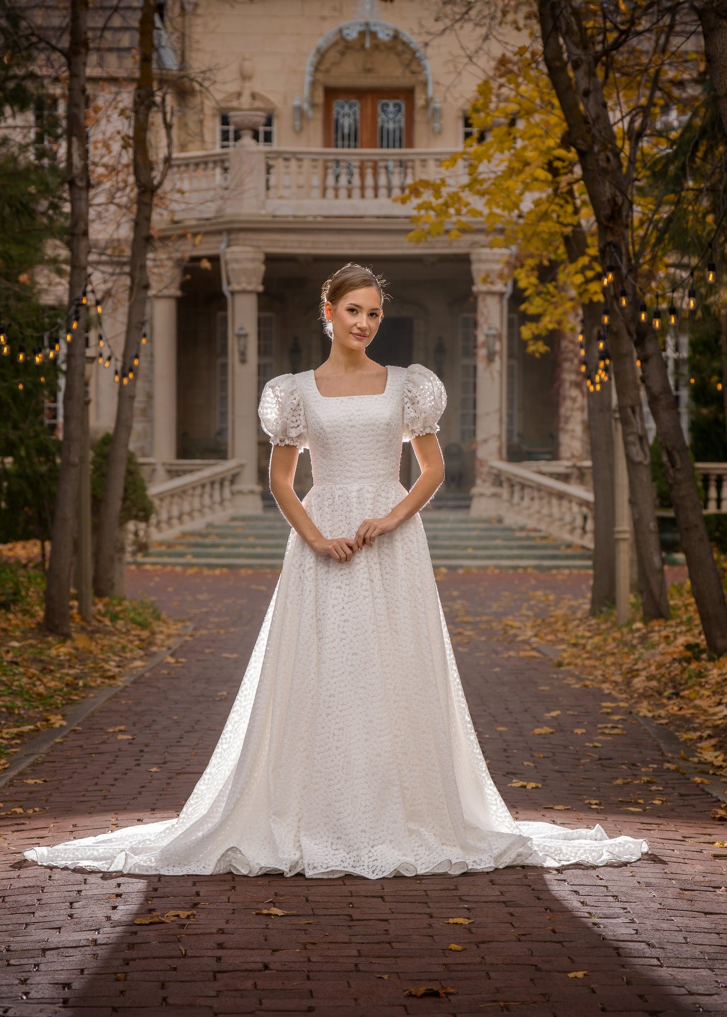 Woman in a white wedding dress standing on a path lined with trees and a building in the background.