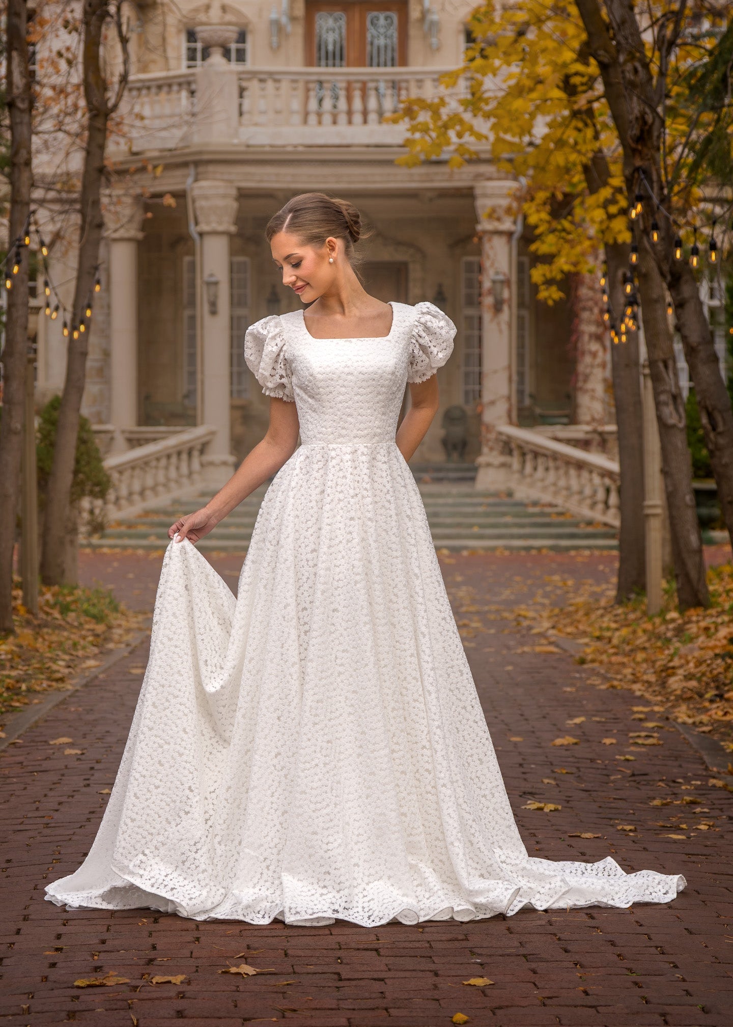 Woman in a white wedding dress standing on a path with trees and a building in the background.
