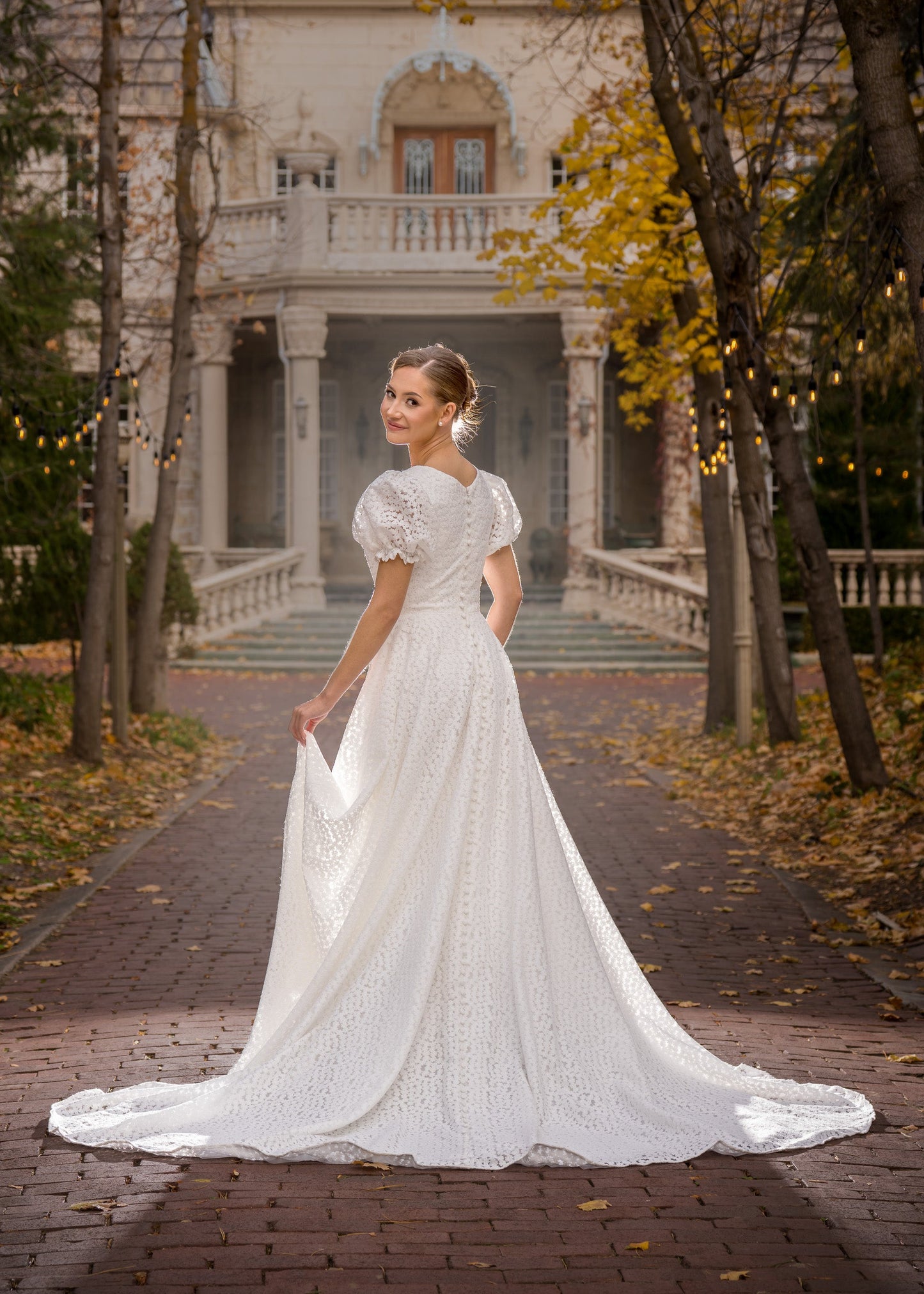 Woman in a white wedding dress standing in front of a large house with autumn foliage.