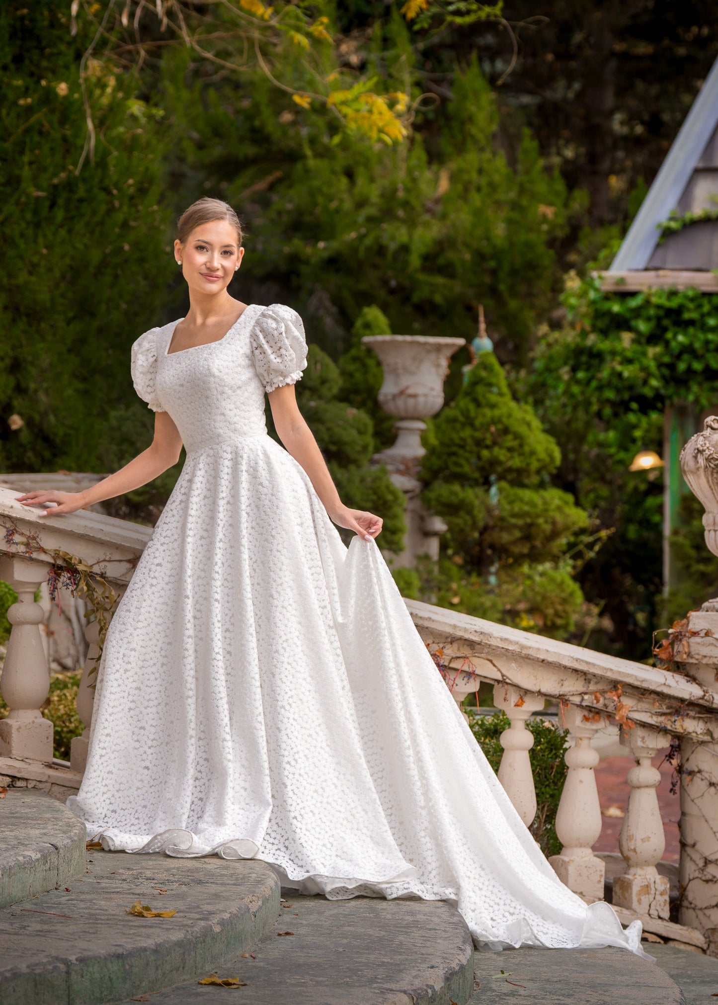 Woman in a white wedding dress standing on a stone pathway with greenery and a building in the background.