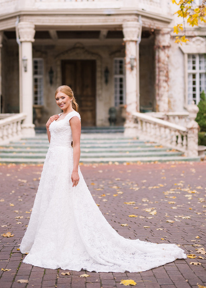 Woman in a white wedding dress standing in front of a large, elegant building with autumn leaves on the ground.