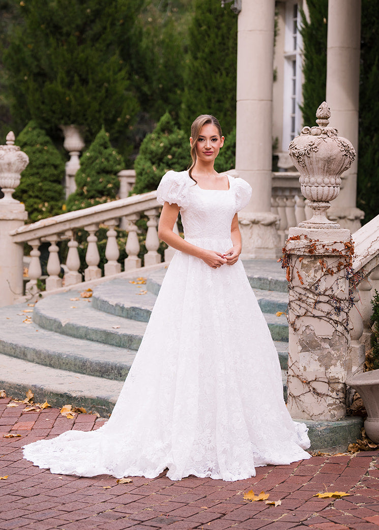Woman in a white wedding dress standing on a brick path with stone columns and greenery in the background.