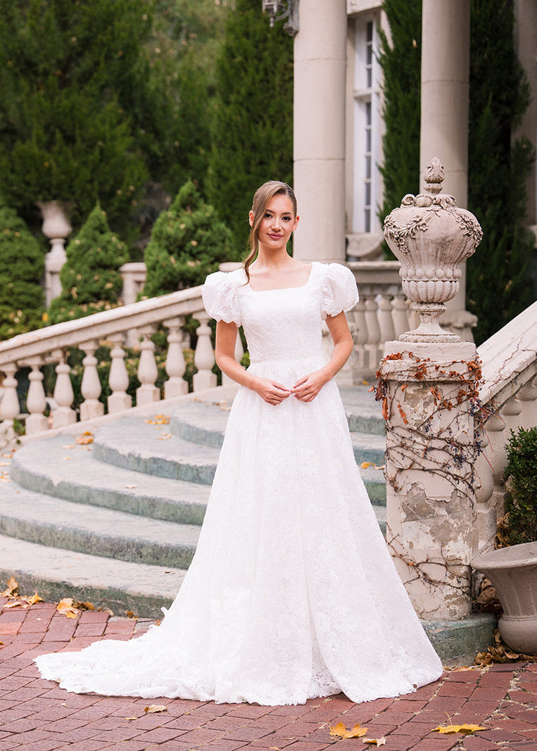 Woman in a white wedding dress standing on a brick path with steps and columns in the background.