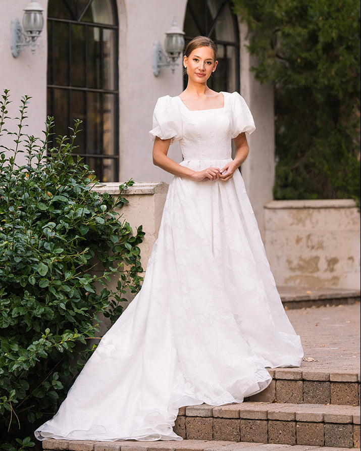 Woman in a white modest wedding dress standing on steps outside a building with greenery.